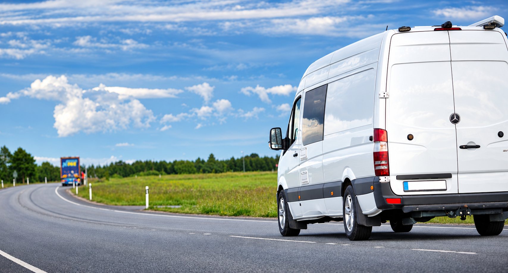 Une camionnette utilitaire blanche de marque Mercedes, vue de l'arrière, circule sur une route de campagne sous un ciel nuageux.