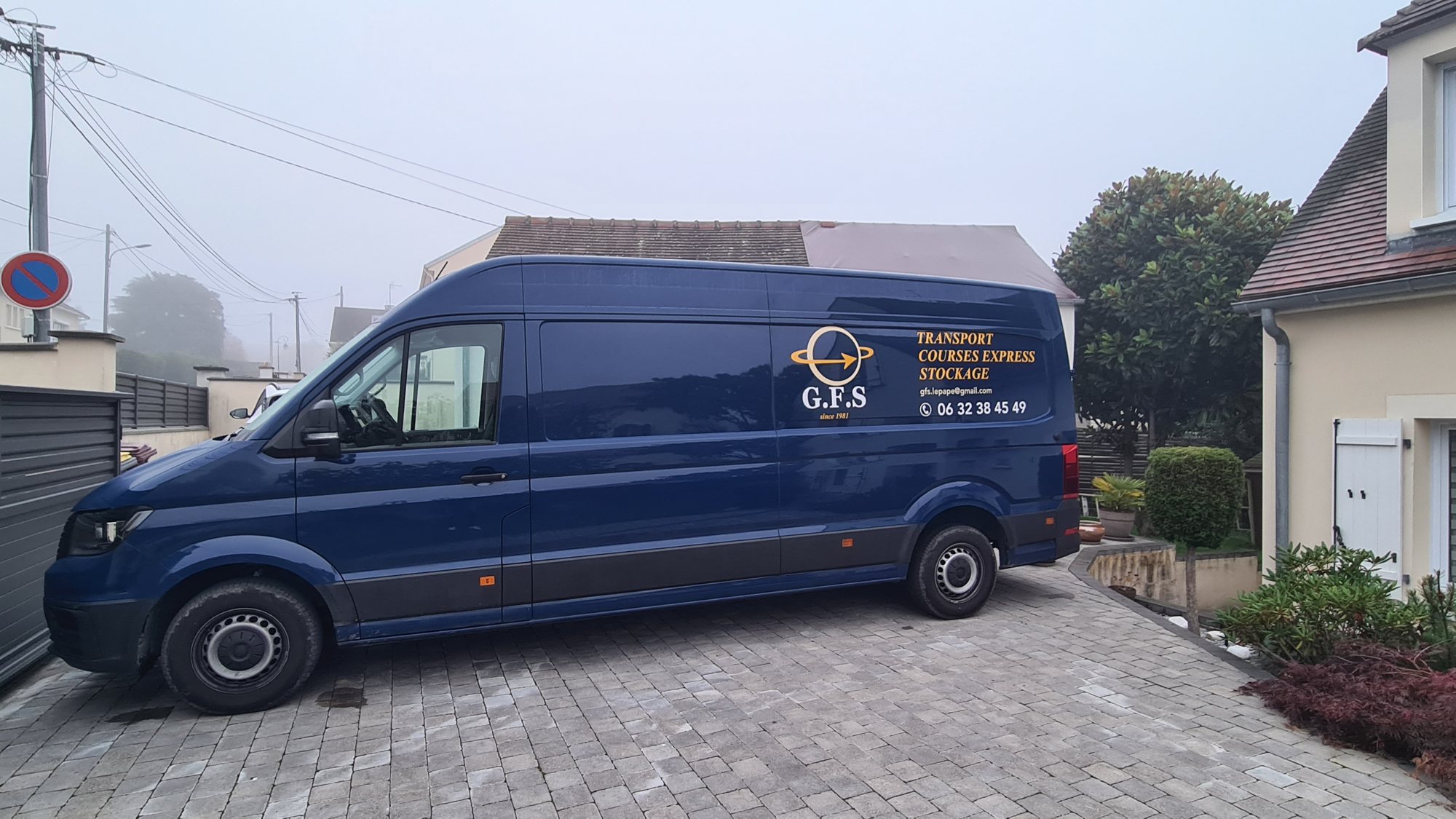 Une grande camionnette bleue portant le logo G.F.S et les mentions transport, courses express et stockage est garée sur une allée pavée par temps de brume.