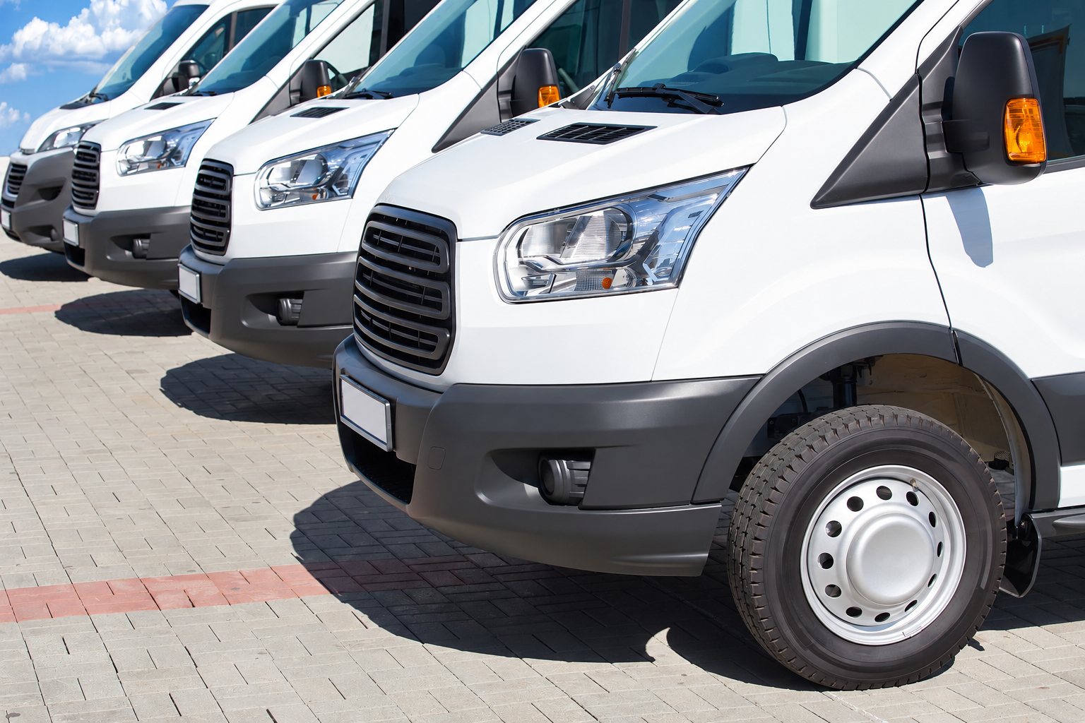 Plusieurs camionnettes blanches sont garées côte à côte sur un sol pavé, sous un ciel bleu.