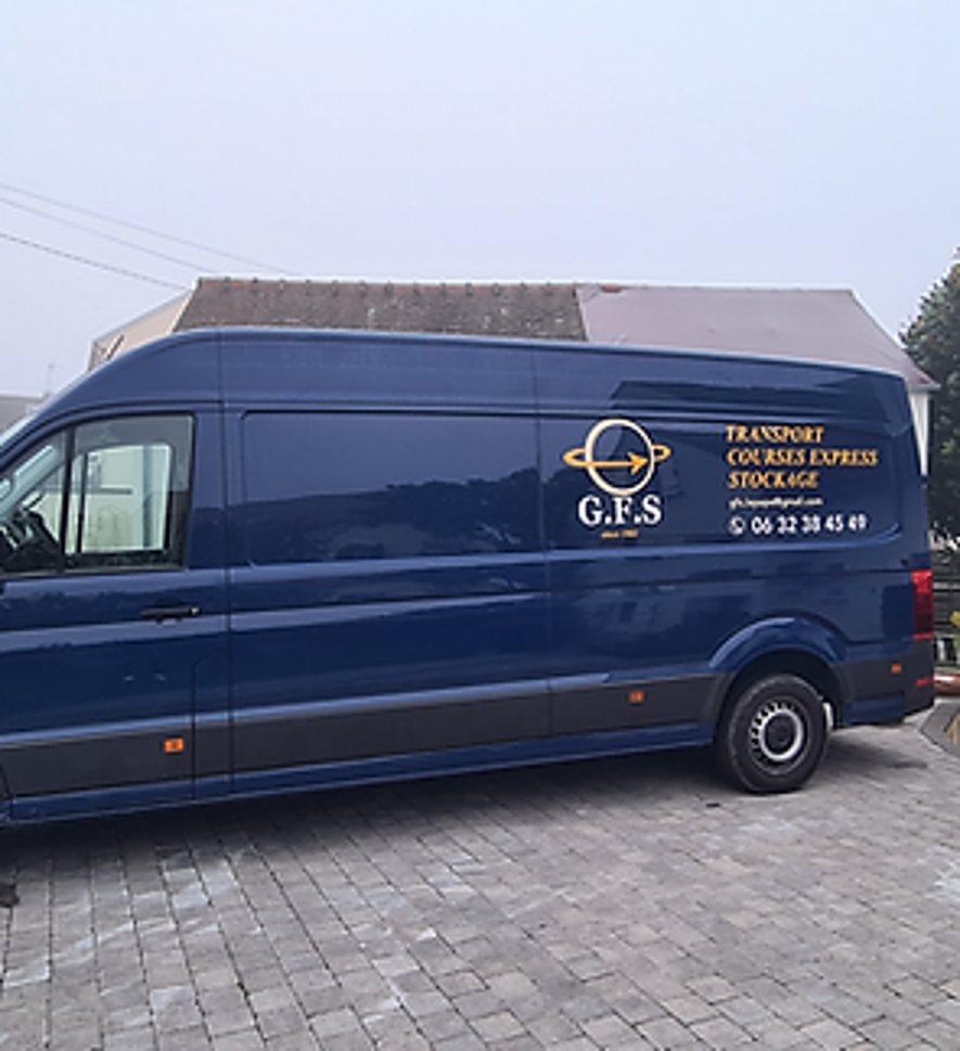 Un grand fourgon utilitaire bleu portant le logo G.F.S et les mentions transport, courses express et stockage, garé sur un sol pavé devant une maison.