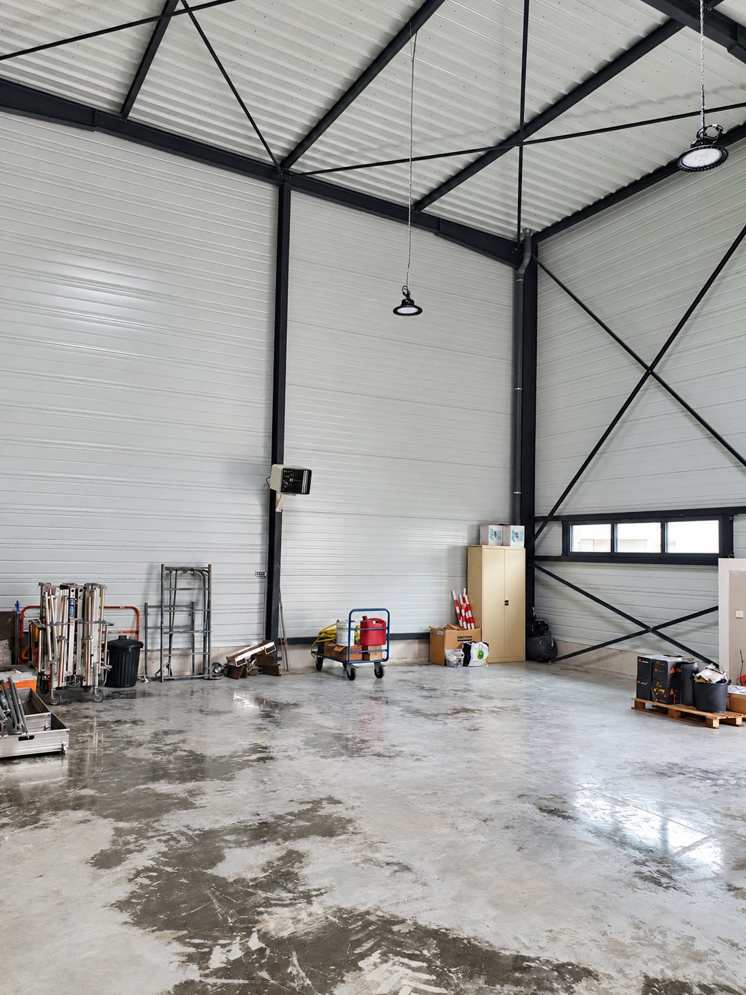 Intérieur d'un grand entrepôt à structure métallique noire, au sol en béton brillant, avec du matériel et une armoire beige rangés le long des murs.