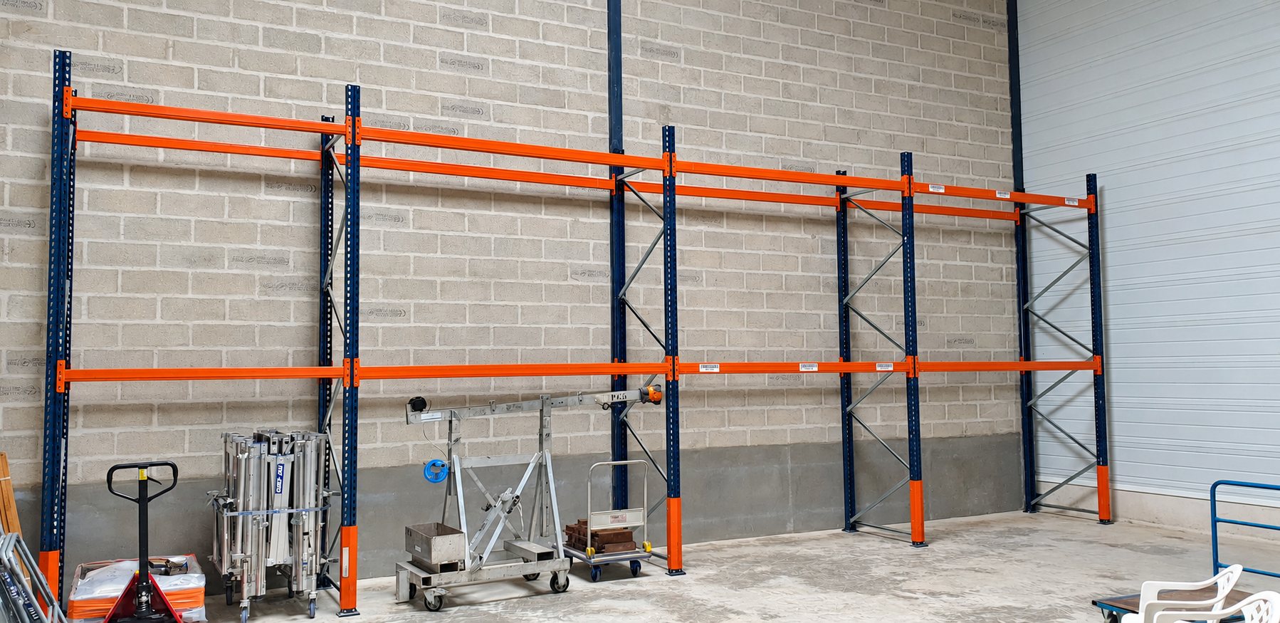 Dans un entrepôt, des rayonnages métalliques orange et bleu vides sont installés contre un mur en parpaings, avec un transpalette et du matériel au sol.